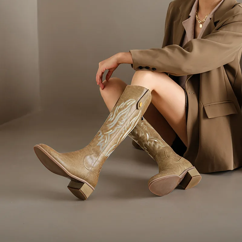 A woman wearing beige chunky-heel western boots with decorative embroidery, paired with a brown blazer