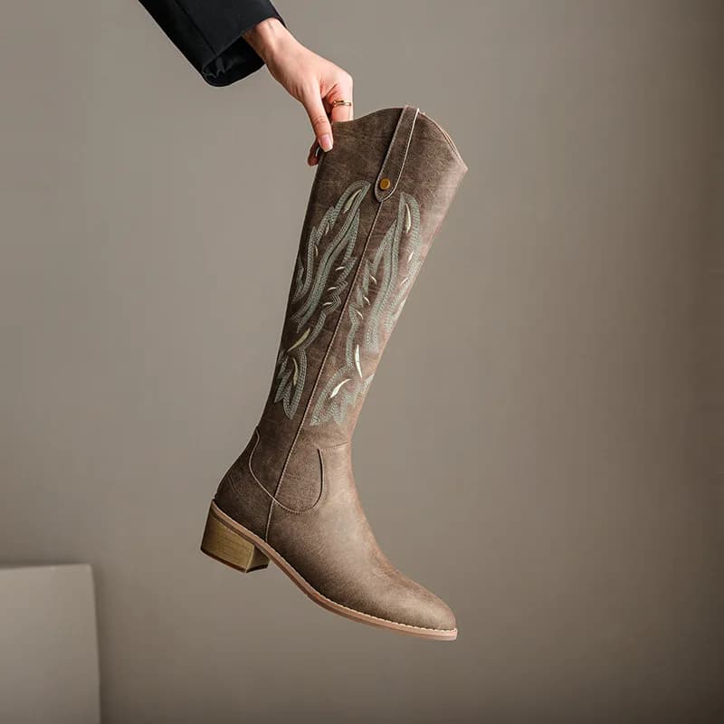 A hand holds up a brown chunky-heel western boot with decorative embroidery against a plain background