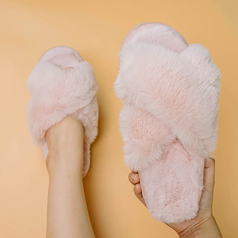 A pair of light pink fuzzy cross-strap slippers, one being worn and one held in a hand