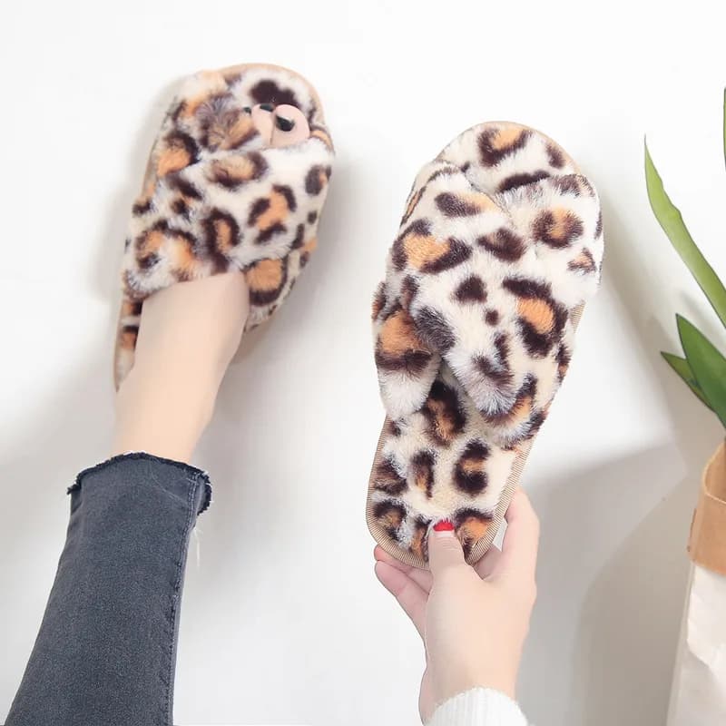 A person wearing one leopard print fuzzy cross-strap slipper and holding the other in their hand