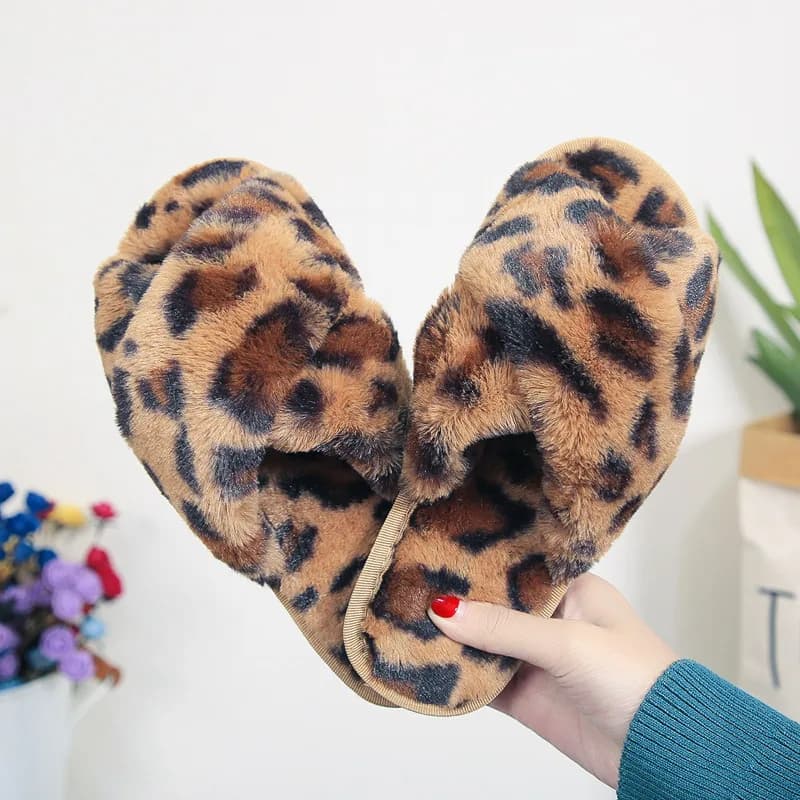A hand holding up a pair of fuzzy leopard print cross-strap slippers against a neutral background