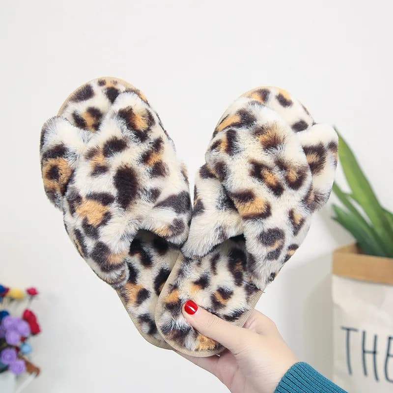 A hand holding up a pair of fuzzy leopard print cross-strap slippers against a neutral background
