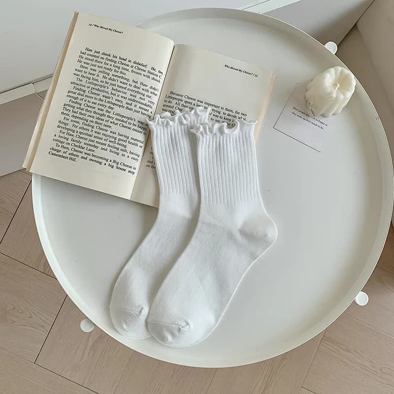 A pair of white ruffle ankle socks resting on a white table next to an open book and a small candle