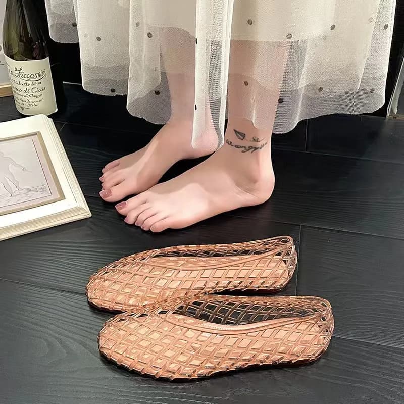 A pair of rose gold woven jelly ballet flats placed on a dark wooden floor near bare feet