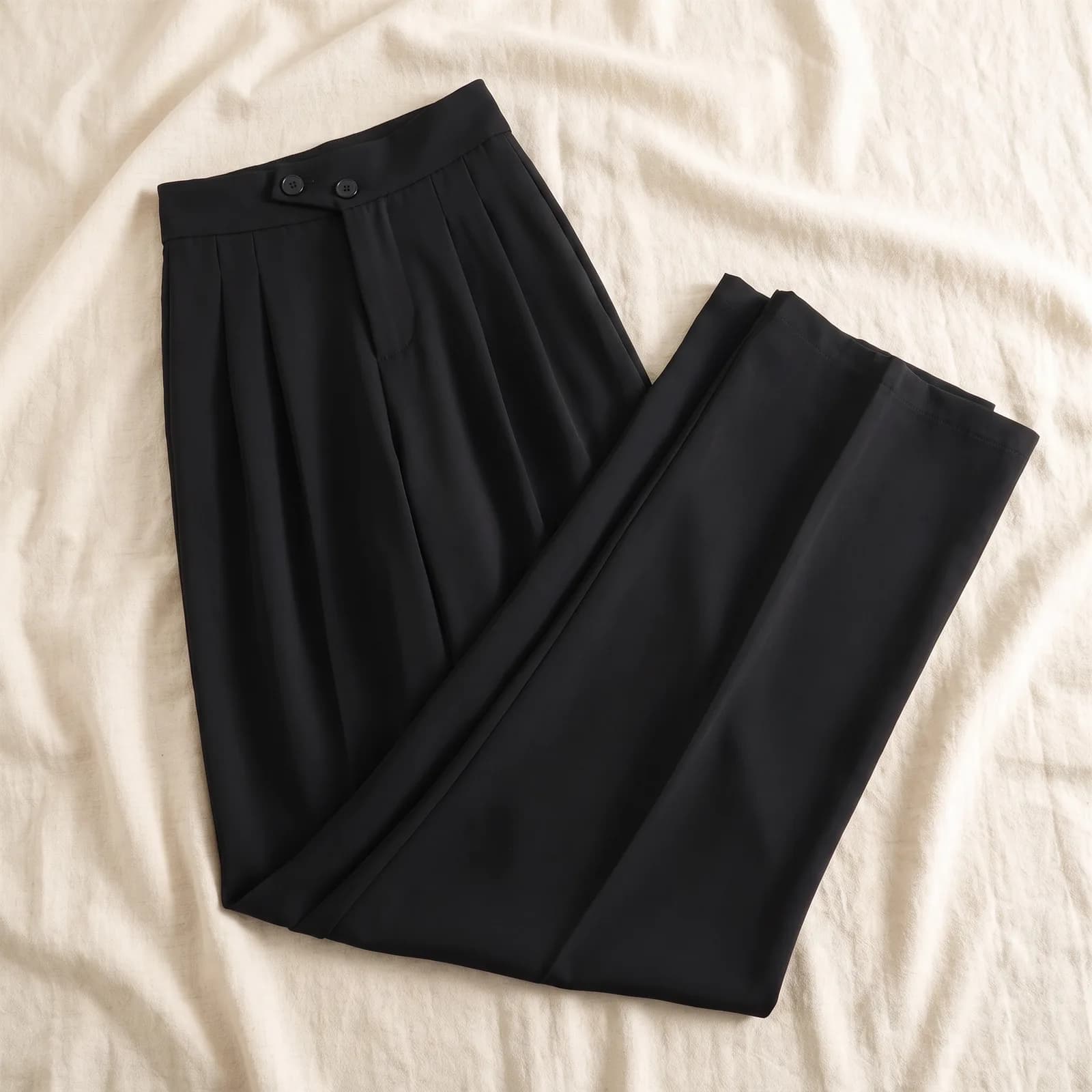 The trousers laid flat top-down on warm cream linen, pleats visible