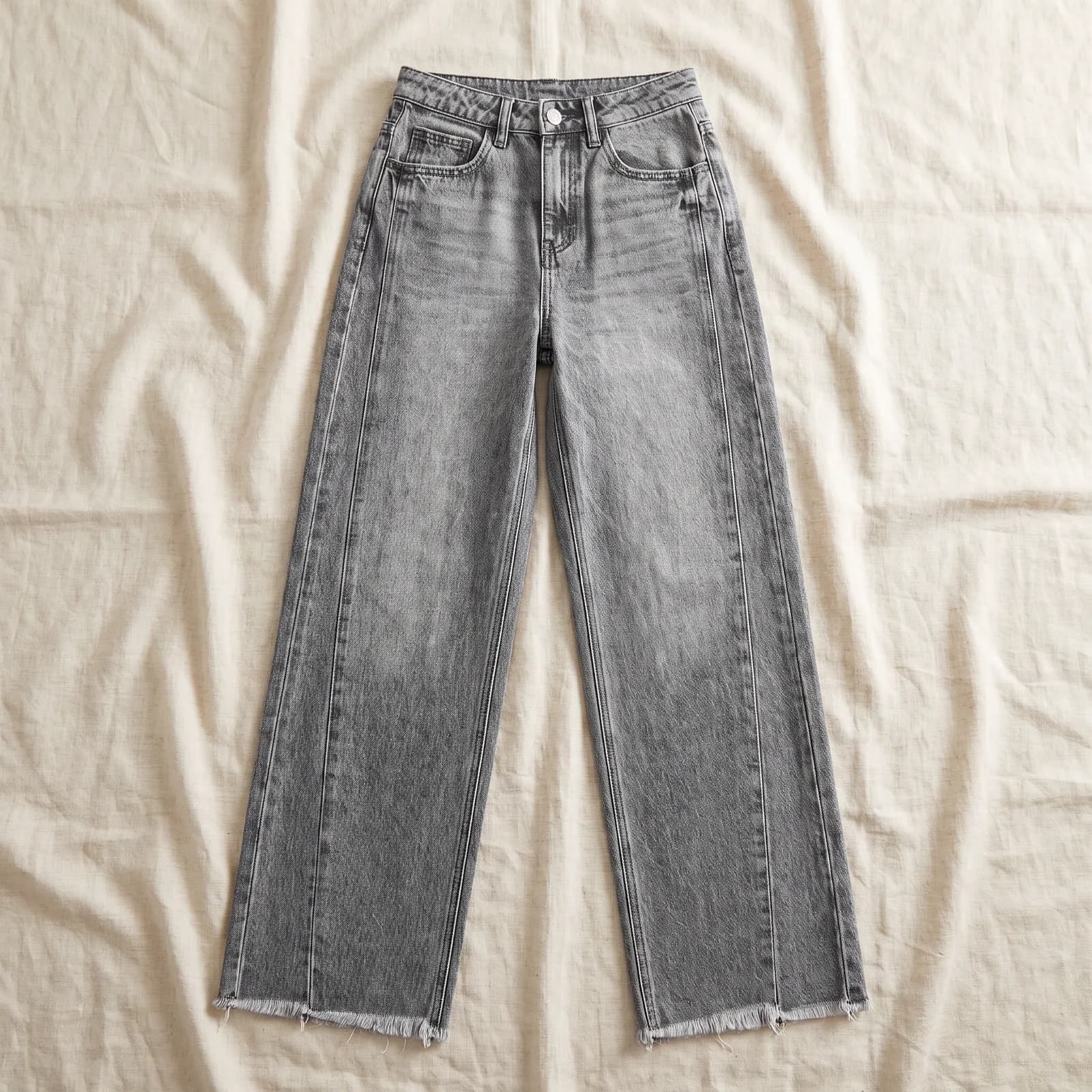 The jeans laid flat top-down on warm cream linen, frayed hem visible