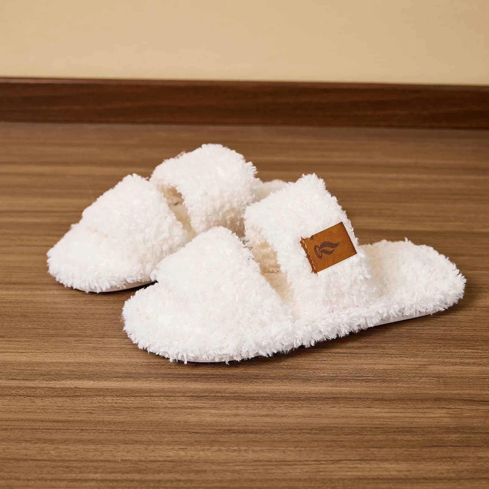 A pair of white plush slide slippers resting on a wooden floor
