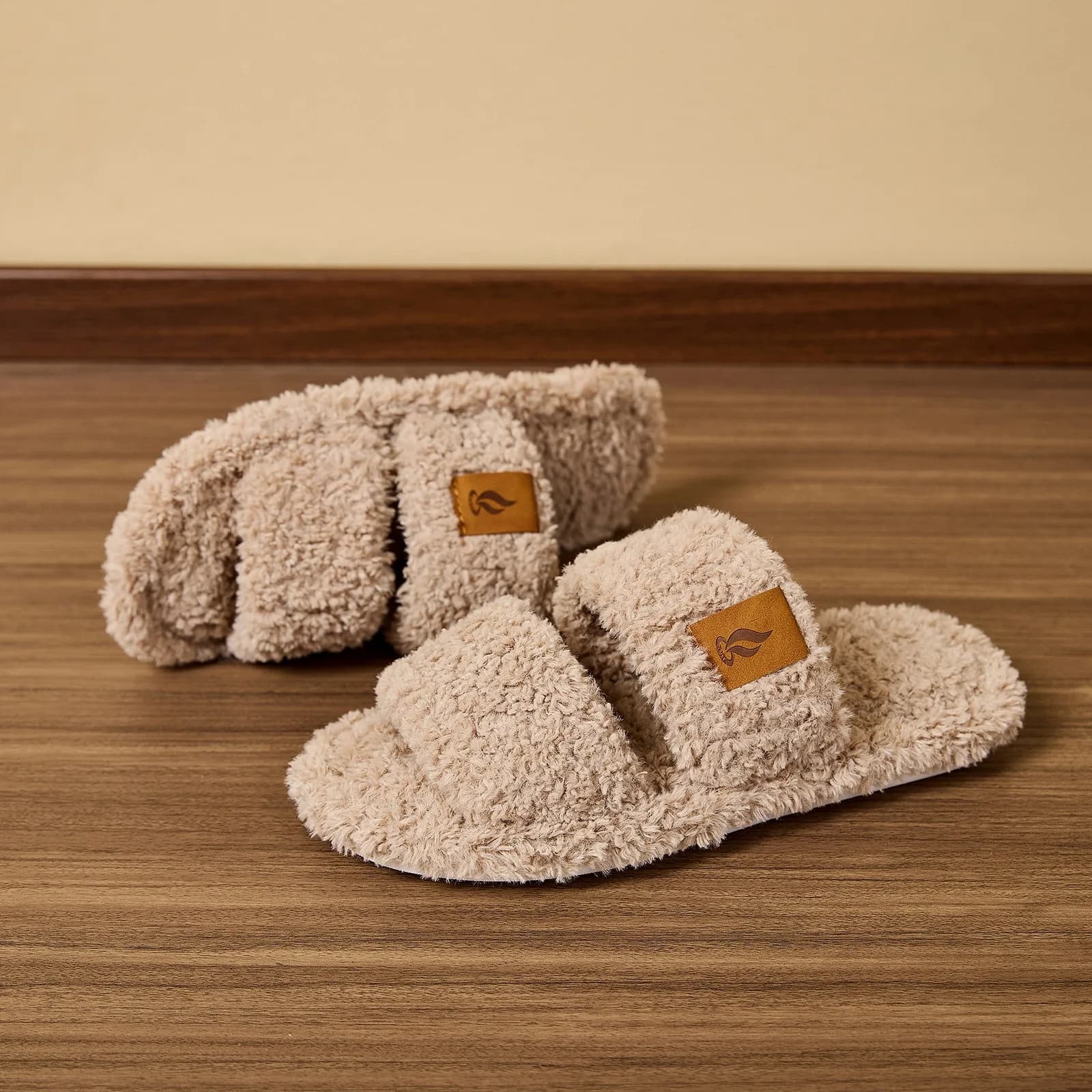 A pair of beige plush slide slippers with a small brown logo tag resting on a wooden floor
