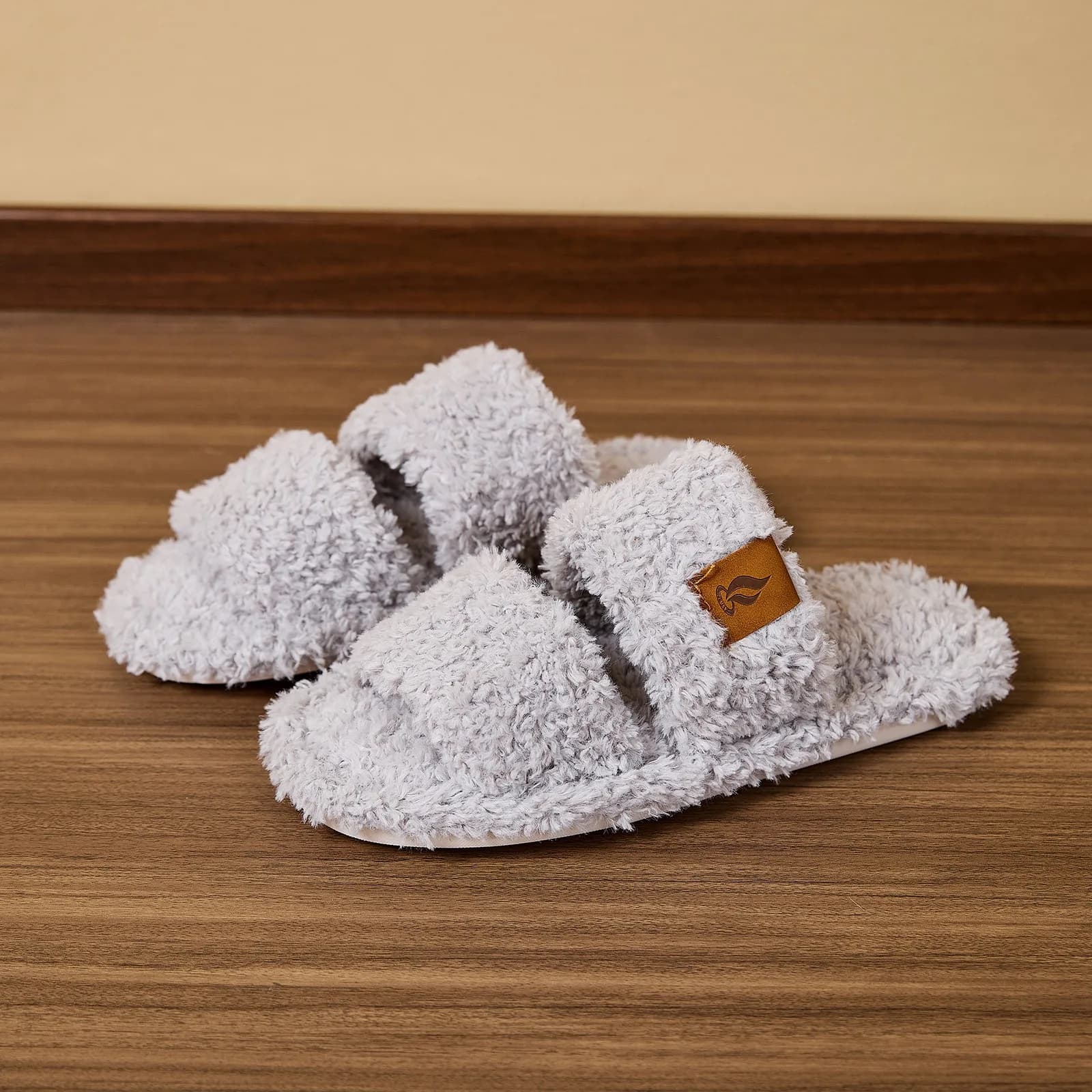 A pair of light grey plush slide slippers resting on a wooden floor