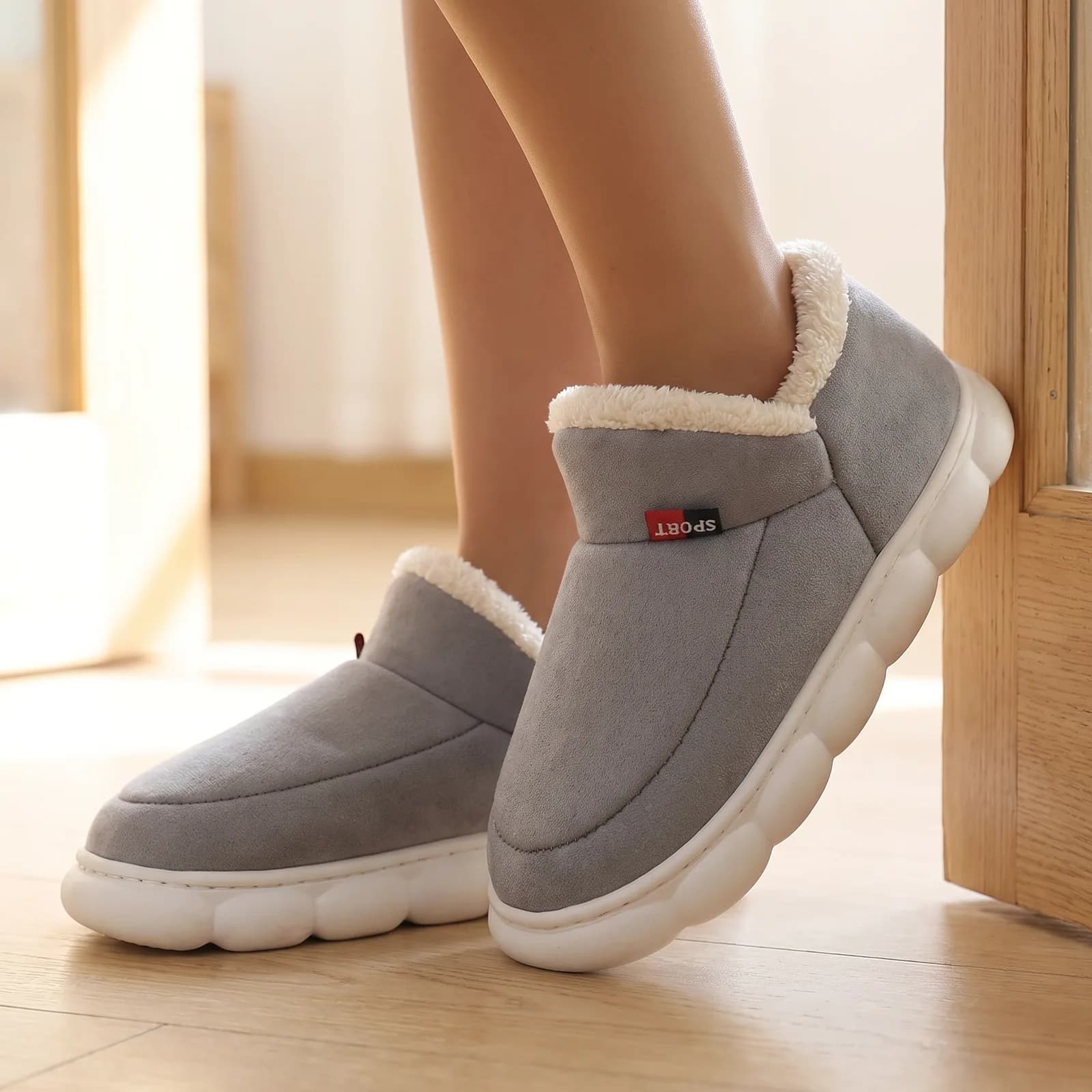 Feet from the ankle down wearing the fleece-lined house shoes on a wood floor